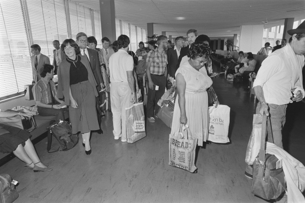 Gruppe von Menschen in einer Schwarz-Weiß-Flughafenszene mit Gepäck, einige sitzen auf Bänken und andere stehen, Fenster mit Jalousien links und ein Mann und eine Frau, die nebeneinander stehen und Taschen halten.