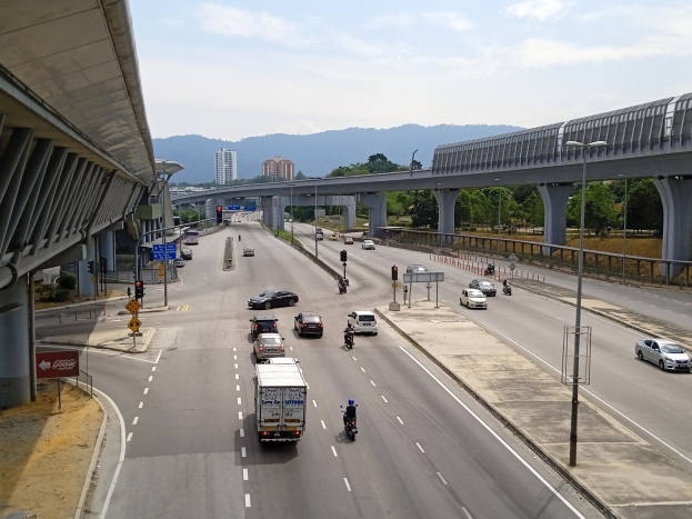 Eine vielbefahrene Autobahn mit mehreren Fahrspuren neben einer Brücke, mit Straßenlaternen, Ampeln, Verkehrsschildern, Bäumen, Gebäuden, Hügeln und einem bewölkten Himmel.