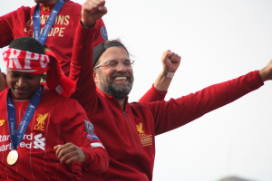 Liverpool-Trainer Jürgen Klopp feiert mit seinen Spielern nach dem Gewinn der UEFA Champions League, während zwei medaillentragende Personen in der Nähe sind und der Himmel im Hintergrund zu sehen ist.