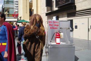 Eine Gruppe von Menschen, einige in Kostümen, versammelte sich vor Gebäuden mit Text darauf, mit einer Box und einem Feuerlöscher im Sichtfeld.