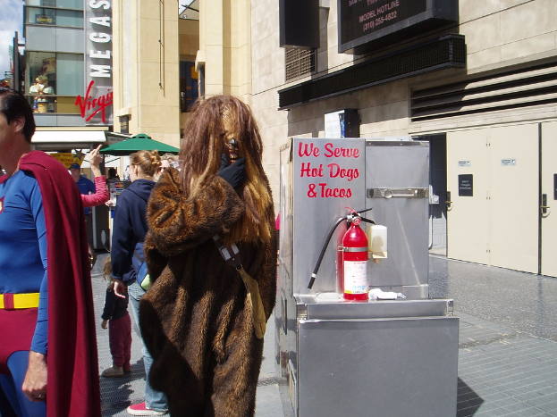 Eine Gruppe von Menschen, einige in Kostümen, versammelte sich vor Gebäuden mit Text darauf, mit einer Box und einem Feuerlöscher im Sichtfeld.