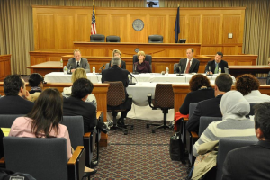 Ein Tagungsraum mit einer Gruppe sitzender Menschen, zwei ausländischen Flaggen, einem orangen Tisch mit Wasserflaschen und einem Mikrofon.