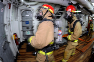 Zwei Feuerwehrleute in Schutzausrüstung, einschließlich Helmen und Gasmasken, gehen durch das Innere eines Flugzeugs, mit einer Tür auf der linken Seite und Rohren und anderen Gegenständen auf der rechten Seite.