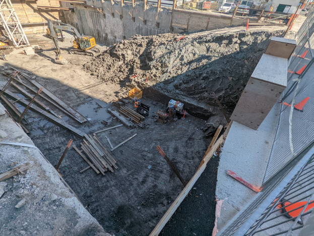 Baustelle mit einem großen Schutthaufen, umgeben von Kränen, Holzstangen und anderem Equipment, abgegrenzt durch einen Maschendrahtzaun, mit Gebäuden, Fahrzeugen, Pfählen, Bäumen und Himmel im Hintergrund.