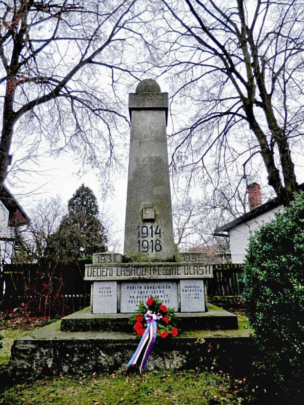 Ein Weltkriegsgedenkmal auf einem Friedhof, geschmückt mit einem Kränzchen, umgeben von Gras und trockenen Blättern, mit Pflanzen, Bäumen, Häusern und Himmel im Hintergrund.