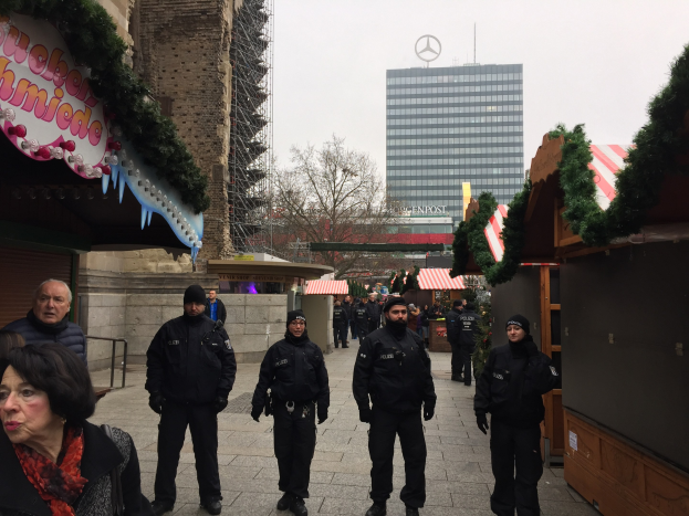 Polizeibeamte in schwarzen Uniformen stehen vor einem geschmückten Weihnachtsmarkt in Berlin, mit festlichen Dekorationen, Gebäuden, Bäumen und einem klaren Himmel im Hintergrund; ein Schild mit Text ist links zu sehen.