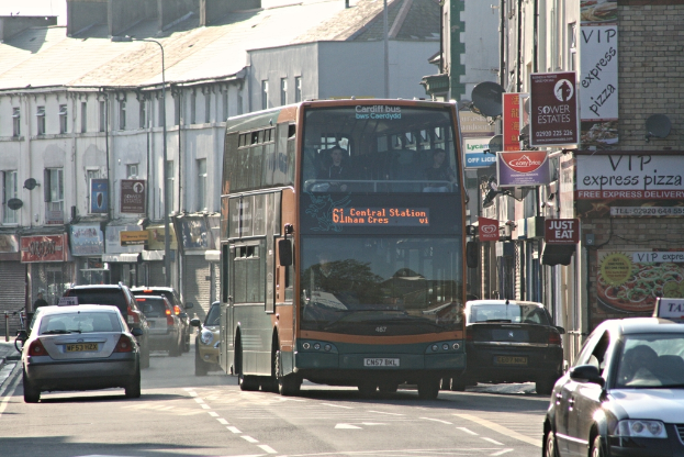 Eine Straße mit Autos und einem Bus vor Gebäuden mit Wänden, Fenstern, Tellern und Dächern, mit Plakaten und Bannern an den Wänden und einem Laternenmast.