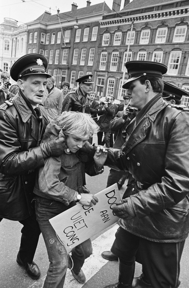 Schwarz-weißes Bild einer Frau, die von zwei Polizeibeamten auf einer Straße eskortiert wird, mit Zuschauern und Gebäuden im Hintergrund.