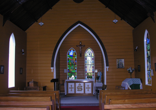 Das Bild zeigt das Innere einer Kirche mit Bänken auf beiden Seiten, einem Kreuz in der Mitte, hängenden Lampen oben, einem Tisch mit einer Vase und einem gerahmten Bild auf der rechten Seite und einem zentralen Idol, das von einem Tisch mit einer Decke flankiert wird.