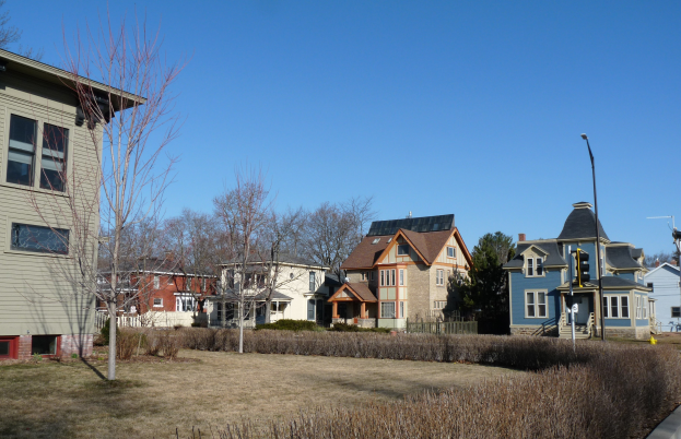 Eine Reihe von Häusern in einem Wohnviertel mit Fenstern, Laternen, Bäumen, Pflanzen, Gras und einem klaren blauen Himmel im Hintergrund.