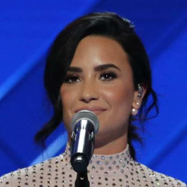 Demi Lovato steht selbstbewusst vor einem Mikrofon bei den Grammys, mit einem schwarzen Kleid und einem eleganten Bob-Haarschnitt, vor einem blauen Hintergrund.