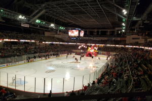 Ein Eishockeyspiel in einer großen Arena mit Zuschauern auf Sitzen und Stehplätzen in der Nähe der Eisfläche, umgeben von Bannern und einem Dach mit Metallstangen, Lampen und zwei Bildschirmen, identifiziert als Columbus Blue Jackets Arena.