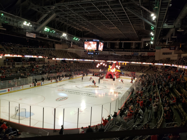 Ein Eishockeyspiel in einer großen Arena mit Zuschauern auf Sitzen und Stehplätzen in der Nähe der Eisfläche, umgeben von Bannern und einem Dach mit Metallstangen, Lampen und zwei Bildschirmen, identifiziert als Columbus Blue Jackets Arena.