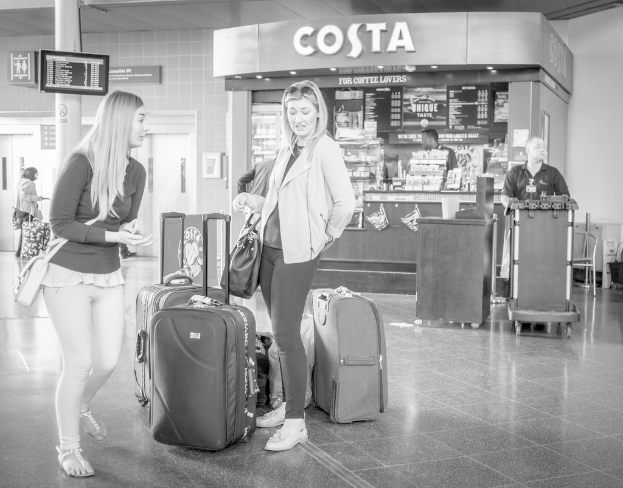 Zwei Frauen vor Gepäckstücken mit einem Kaffee-Shop im Hintergrund, wahrscheinlich am Flughafen.