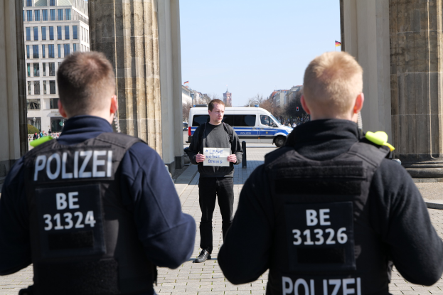 Drei Polizeibeamte stehen vor einem Gebäude, wobei ein Mann in der Mitte ein Papier hält, flankiert von Säulen und einer belebten städtischen Umgebung.