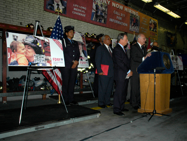 Eine Gruppe von Menschen steht in der Nähe eines Podiums mit Mikrofonen, umgeben von Tafeln auf Ständern, Fahnen und Tafeln an der Wand, alles unter hellen Lichtern.
