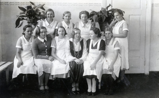Eine Gruppe von Frauen, die auf einer Bank in einem Raum mit einer Wand, einer Tür und Zimmerpflanzen im Hintergrund sitzen.