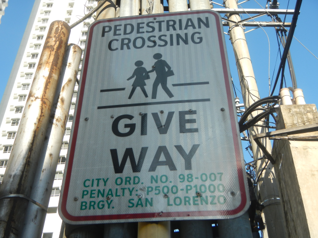 Ein Mast mit einer Tafel, auf der "Fußgängerübergang Vorrang" steht, umgeben von Strommasten und -leitungen, im Hintergrund ein Gebäude und der Himmel.
