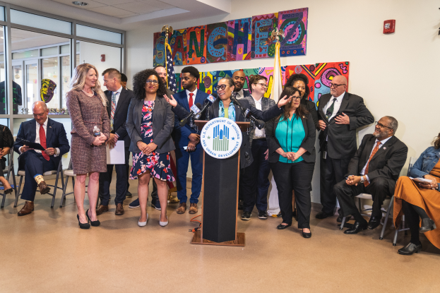 Eine Gruppe von Menschen, die vor einem Rednerpult mit Mikrofon und einer Tafel mit Text stehen und sitzen, mit Flaggen, einer Tafel, einem Feuerlöscher und Deckenleuchten an der Rückwand.