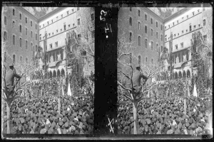 Ein Schwarz-Weiß-Foto einer großen Menschenmenge vor einem Gebäude mit Fenstern, Bäumen und Flaggen im Hintergrund, die scheinbar eine Protestveranstaltung abhalten, wobei einige Schilder und Banner tragen.