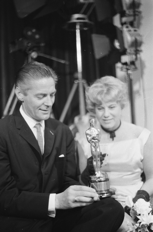 Ein Mann in einem schwarzen Blazer, weißem Hemd und Krawatte hält eine Oscar-Statue in der Hand und sitzt neben einer Frau, die einen Blumenstrauß hält, beide in eleganter Kleidung, mit abgedunkeltem Hintergrund, schwarz-weiß.