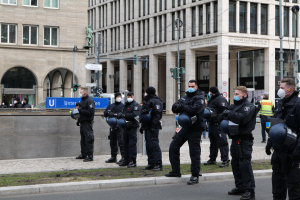 Eine Gruppe von Polizisten in schwarzen Uniformen und Masken steht vor einem Berliner Gebäude mit Glasfenstern, Säulen und umliegenden Elementen wie Laternen, Ampeln und einer Statue.