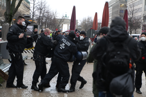 Eine Gruppe von Polizisten in schwarzer Einsatzkleidung und Masken geht eine Straße in Berlin, Deutschland, entlang, mit Gebäuden, Bäumen, Schirmen, Pfosten, Fahrzeugen und einem sichtbaren Himmel im Hintergrund.