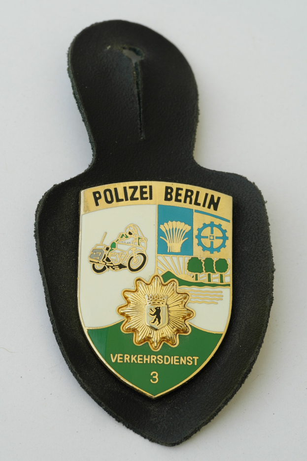 Ein schildförmiges Polizeibadge mit 'Polizei Berlin' in schwarzer Schrift auf einem blauen Hintergrund, das ein weißes Stern und einen weißen Rand hat und auf einer Oberfläche liegt.