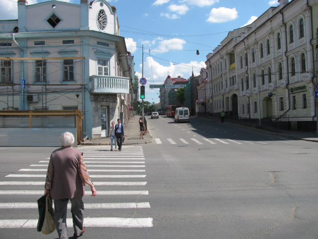 Alte Frau überquert eine Straße vor einem Gebäude mit Fußgängern, Fahrzeugen, Schildern, Strommasten, Bäumen und einem bewölkten Himmel im Hintergrund.