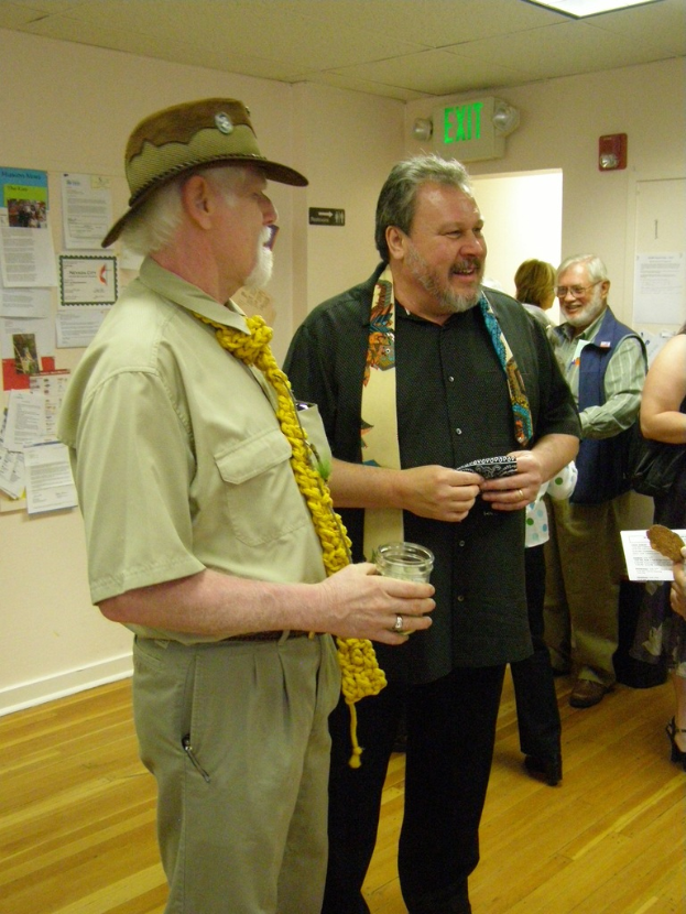 Eine Gruppe älterer Männer steht auf einem Holzfußboden in einem Raum mit Papieren an den Wänden, einer Warntafel an der rechten Wand, einer Tür darunter und Deckenlampen.