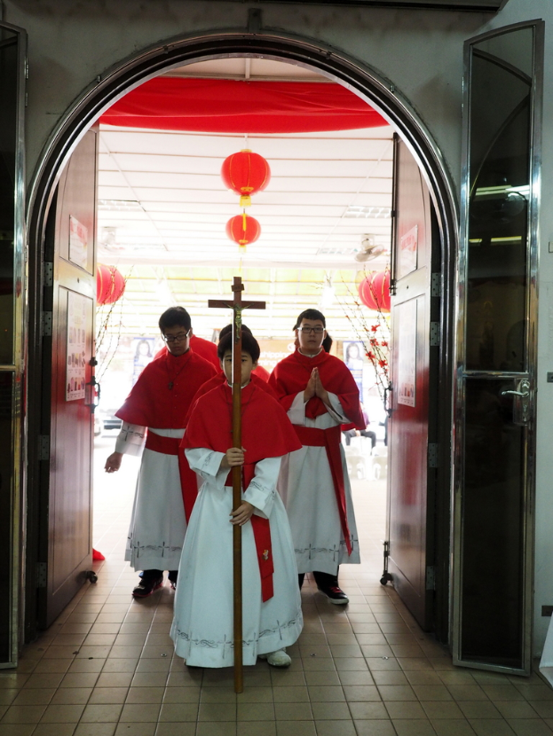 Menschen mit einem Kreuz in einer Kathedrale, mit Türen links und rechts und Blumen und Dekorationen im Hintergrund.