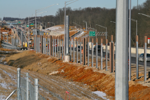 Baustelle mit Fahrzeugen, Pfählen, Lichtern, Beschilderung, einem Zaun, schneebedecktem Gras, Bäumen und einem Himmel im Hintergrund.