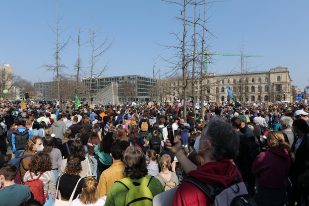 Große Menschenmenge mit Schildern und Taschen vor einem Gebäude mit Bäumen und klarem Himmel, was auf eine Klimaprotestveranstaltung hinweist.