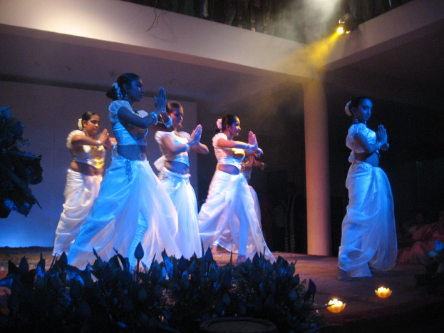 Eine Gruppe von Menschen tanzt auf einer Bühne mit Pflanzen davor und einem Publikum, das zuschaut.