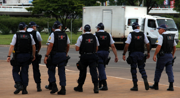 Eine Gruppe von Uniformierten, darunter Mützen, Hemden, kugelsichere Westen, Uhren, Hosen und Schuhe, die auf einer Straße mit einem geparkten Lkw auf der rechten Seite neben einem Zaun gehen. Im Hintergrund sind Gebäude, Bäume, Pflanzen, Pfosten, Banner und eine Brücke oben rechts zu sehen.