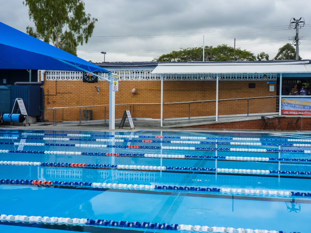 Ein großes Schwimmbad mit Bahnentrennern, umgeben von einem Zaun, einem Schuppen, einer Fahne, Stühlen, einem Tisch, einem Schild, Pfosten, einem Straßenpfosten, Drähten, Bäumen und einem bewölkten Himmel.