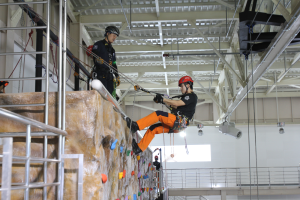 Zwei Personen in schwarzen und orangen Outfits und Helmen klettern an einer Felswand in einer Halle, gesichert durch Seile, mit einem Metallgeländer links und einer weißen Wand mit Lampen im Hintergrund.