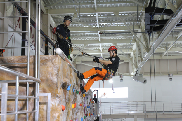 Zwei Personen in schwarzen und orangen Outfits und Helmen klettern an einer Felswand in einer Halle, gesichert durch Seile, mit einem Metallgeländer links und einer weißen Wand mit Lampen im Hintergrund.