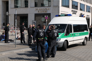 Eine Gruppe von Polizisten steht vor einem Starbucks-Café, rechts daneben parkt ein Van, links stehen und sitzen Menschen, im Hintergrund ein Gebäude mit Fenstern, ein Schild, ein Laternenmast und ein Zaun.