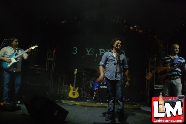 Drei Personen auf der Bühne, eine spielt Gitarre und eine andere singt in ein Mikrofon, mit einer Snare-Drum-Anordnung und Beleuchtung im Hintergrund.