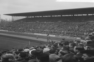 Ein Schwarz-Weiß-Foto eines vollen Stadions mit Menschen, die ein Fußballspiel angucken, Bannern, Pfählen, einer Hütte, Bäumen, einem Turm und einem bewölkten Himmel im Hintergrund.