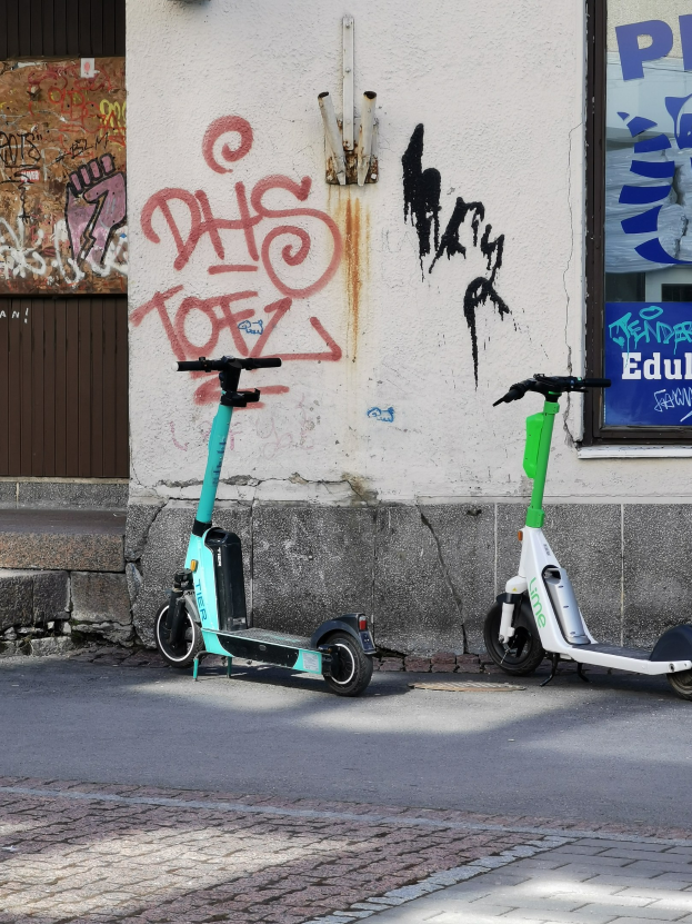 Zwei Elektro-Scooter nebeneinander auf einer Straße geparkt, mit einer Graffiti-bedeckten Wand und einem Plakat im Fenster im Hintergrund.