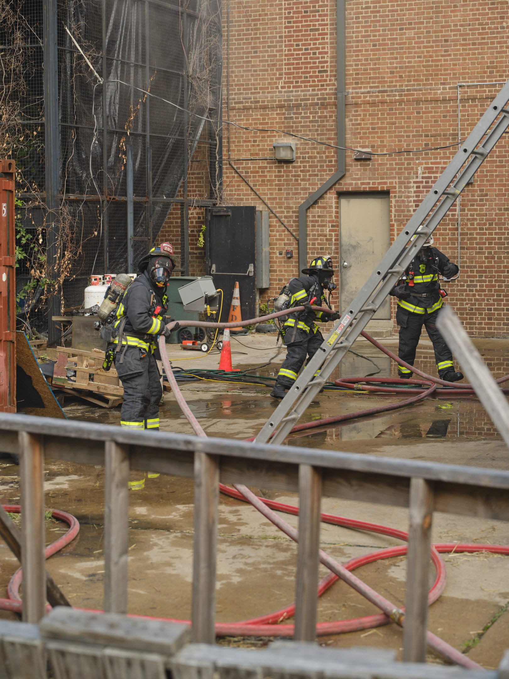 Feuerwehrleute in Helmen arbeiten daran, ein Gebäude Feuer zu löschen, umgeben von Ausrüstung und einem Metallzaun, mit einem Baum und Himmel im Hintergrund.