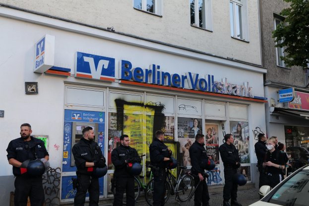 Eine Gruppe von Polizeibeamten vor einer Berliner Volksbank, einige halten Helme in den Händen, mit einem Auto auf der rechten Seite und einem Baum auf der linken Seite sowie Gebäuden mit Fenstern und Texttafeln im Hintergrund.