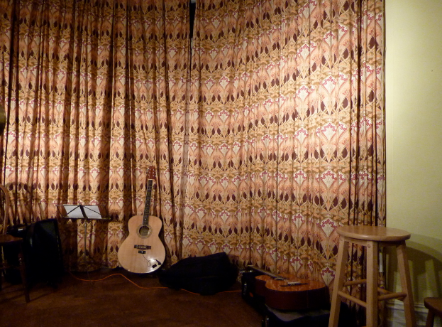 Hocker, Gitarren, ein Buch auf einem musikalischen Buchständer und verschiedene Gegenstände im Vordergrund mit Vorhängen und einer Wand im Hintergrund.
