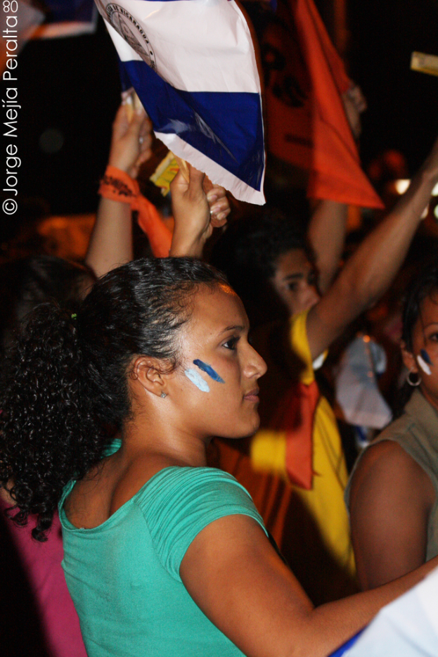 Eine Frau in einem grünen T-Shirt steht bei einer Protestaktion und hölt ein weißes Schild, hinter ihr stehen viele andere Menschen.