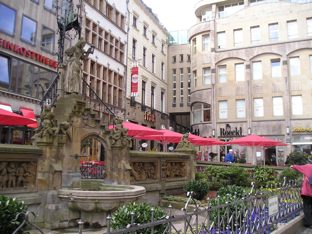 Eine Stadtlandschaft mit Gebäuden, Zelten, Pflanzen und Menschen im Hintergrund und einer Skulptur, einem Teich, einem Geländer und Pflanzen im Vordergrund.