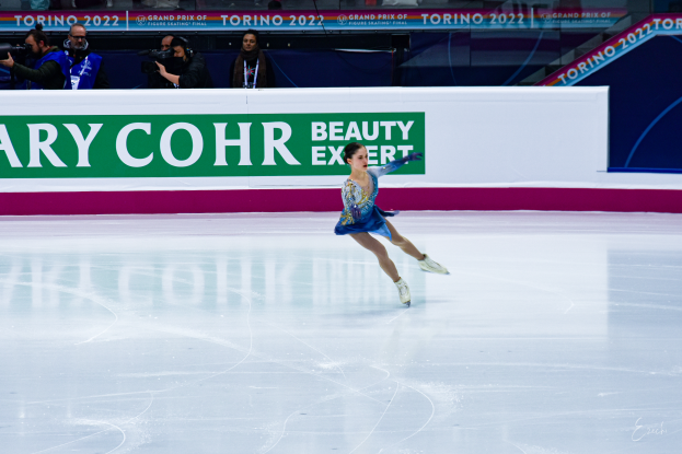 Eine Frau in einem blauen Kleid läuft auf einem Eisstadion während eines professionellen Eiskunstlauf-Events, umgeben von einer Menge mit Kameras und einem Schild mit der Aufschrift "Tessa Virtue und Scott Moir" im Hintergrund.