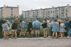 Eine Gruppe von Kindern steht vor einem Gebäude, umgeben von blühenden Pflanzen und Bäumen, unter einem klaren blauen Himmel, mit einigen, die Taschen halten.