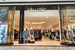 Ein stylischer Mode-Boutique in einem Einkaufszentrum, mit Schaufensterpuppen in der neuesten Mode, Kleidung an Bügeln, einer Säule, einem Schild mit Text, Deckenleuchten und einem Fliesenboden.